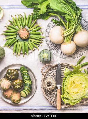 Verdure verdi in ciotole sul tavolo luminoso con coltello: piselli verdi, cavolo rapa, lattuga, zucchini, cetrioli, pomodori verdi. Vista dall'alto. Pulire mangiare vegetariano Foto Stock