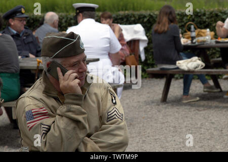 Un WW2 rievocazione storica di un esercito americano sergente di 101st Airborne in camicia di Ike e tappo di guarnigione si rilassa su un giorno moderno telefono cellulare Foto Stock