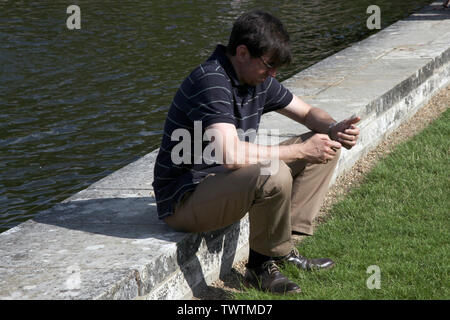 Edenbridge, Kent, Regno Unito - Uomo seduto accanto al lago tramite telefono cellulare durante il giorno sun Foto Stock