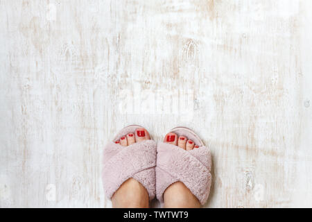 Gambe femmina rosso con chiodi in home pellicce soffici pantofole rosa su una luce sullo sfondo di legno. flat laici. Vista dall'alto. Il concetto di un accogliente luminosa casa ragazza. Foto Stock