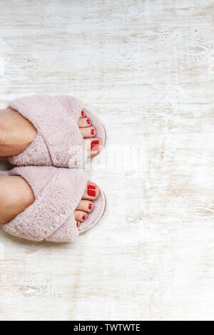 Gambe femmina rosso con chiodi in home pellicce soffici pantofole rosa su una luce sullo sfondo di legno. flat laici. Vista dall'alto. Il concetto di un accogliente luminosa casa ragazza. Foto Stock