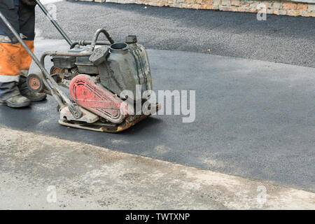 I lavoratori stradali compact l'asfalto con una vibrante rammer sulla strada. Lavoratore con compattatore a una strada sito in costruzione. Foto Stock