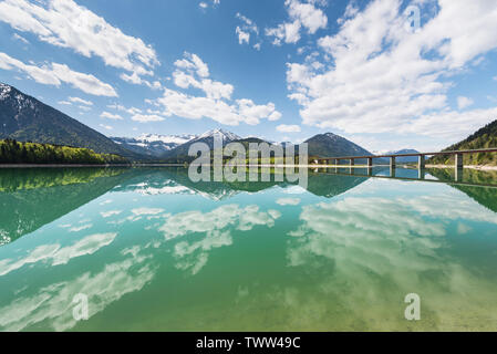 Il cielo blu con nuvole bianche sopra il serbatoio Sylvenstein di fronte montagne innevate di Karwendel con riflessi nell'acqua turchese, Bavaria Foto Stock