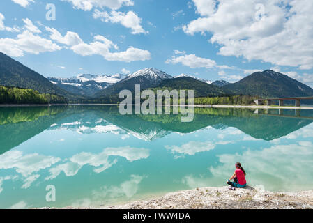 Donna seduta sulla riva del serbatoio Sylvenstein e guardando le montagne Karwendel riflessa nell'acqua turchese, Baviera, Germania Foto Stock