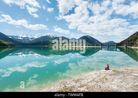 Donna seduta sulla riva del serbatoio Sylvenstein e guardando le montagne Karwendel riflessa nell'acqua turchese, Baviera, Germania Foto Stock