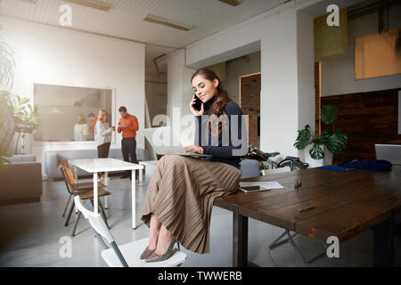 A piena lunghezza shot di giovane imprenditrice parlando con qualcuno sul suo telefono cellulare mentre è seduto in ufficio e di lavoro. Foto Stock
