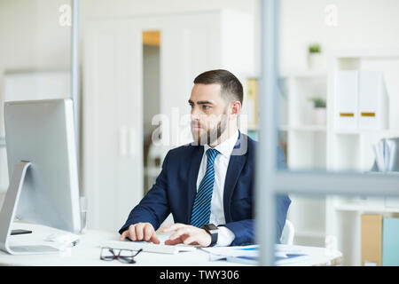Considerato grave giovane imprenditore barbuto in blu scuro tuta seduta a tavola e digitando sulla tastiera del computer mentre si analizzano i dati statistici Foto Stock