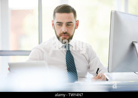 Grave concentrato barbuto giovani manager in camicia formale seduto alla scrivania in ufficio e lavora sul progetto utilizzando i moderni dispositivi Foto Stock