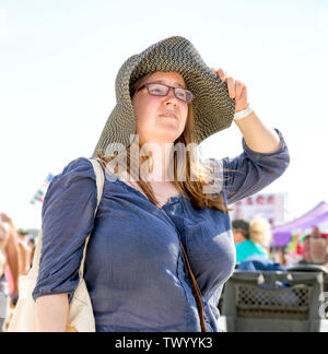 Una giovane donna bruna con gli occhiali tiene il suo cappello dall'orlo. Texas Sandfest 2019 in Port Aransas, Texas, Stati Uniti d'America. Foto Stock