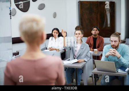 Ampio angolo ritratto contemporaneo di giovane donna alzando la mano in classe con gruppo di studenti godendo di una lezione, insegnanti punto di vista Foto Stock