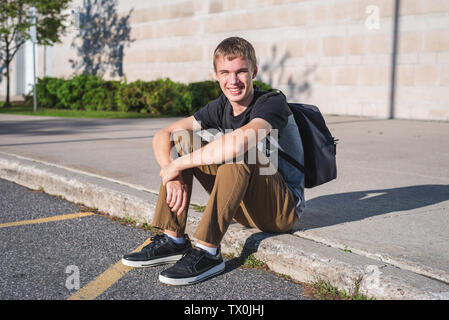 Felice ragazzo seduto sul marciapiede davanti di alta scuola. Foto Stock