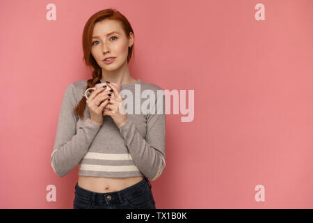 Redhead giovane donna detiene una tazza e guarda la telecamera su sfondo rosa Foto Stock