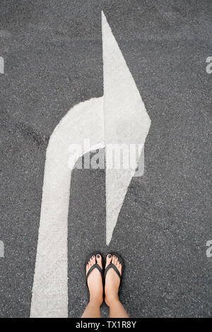 Girare a destra simbolo del traffico. I piedi e le frecce sulla strada dello sfondo. Donna nero scarpe o sandali con il nero smalto per unghie Manicure piedi sulla strada bianca. Foto Stock