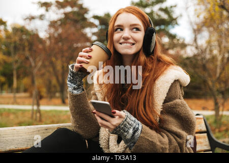Redheaded graziosa fanciulla l'ascolto di musica con le cuffie mentre è seduto su un banco di lavoro, usando il telefono cellulare Foto Stock