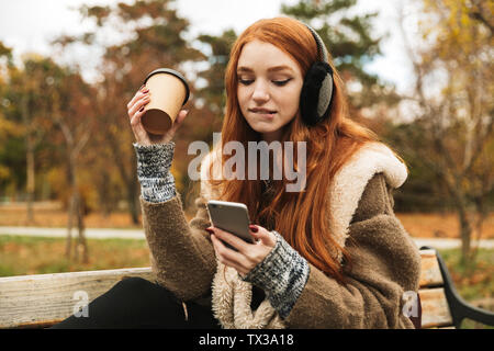 Redheaded graziosa fanciulla l'ascolto di musica con le cuffie mentre è seduto su un banco di lavoro, usando il telefono cellulare Foto Stock