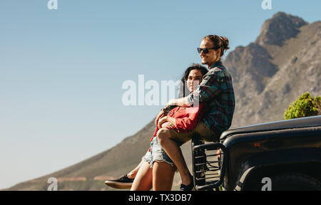 Uomo seduto sul cofano per auto e abbracciando la sua ragazza. Amare giovane su un roadtrip. Foto Stock