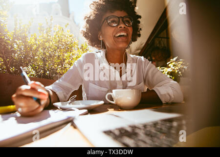 Felice giovane donna africana la scrittura di note durante la seduta a una caffetteria con un computer portatile sul tavolo. Lavoro femminile da un coffee shop. Foto Stock