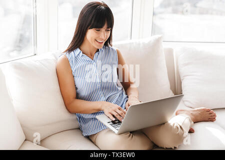 Ritratto di donna sorridente e la digitazione utilizzando argento portatile mentre è seduto sul lettino in luminoso appartamento Foto Stock
