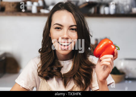 Allegro giovane ragazza che indossa il grembiule seduta in cucina a casa, tenendo cupsicum Foto Stock