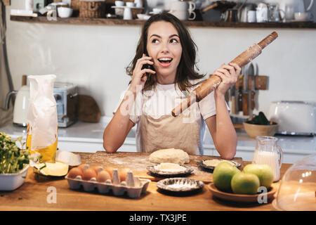 Allegro giovane donna indossando grembiuli preparare la pasta per una fetta di torta di mele in cucina a casa utilizzando il telefono cellulare, trattenendo il perno di rotolamento Foto Stock