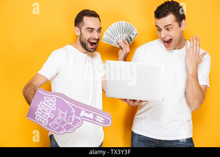 Due allegro eccitato gli uomini amici indossando fustellato t-shirt in piedi isolato su sfondo giallo, utilizzando il computer portatile, celebrando il successo, mostra Foto Stock