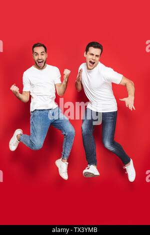 Lunghezza completa di due allegro eccitato gli uomini amici indossando fustellato t-shirt jumping isolate su sfondo rosso, celebrando Foto Stock