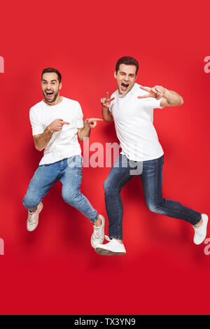 Lunghezza completa di due allegro eccitato gli uomini amici indossando fustellato t-shirt jumping isolate su sfondo rosso, celebrando Foto Stock