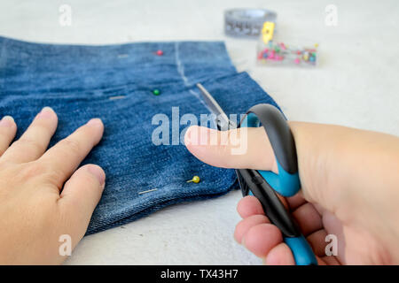 Donna mano che tiene una forbice e taglio di un blue jeans corti a renderli più corti. Rendendo il denim shorts. Su uno sfondo bianco. Foto Stock