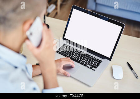 Giovane uomo parlando al telefono durante la digitazione su computer portatile in ufficio in casa Foto Stock