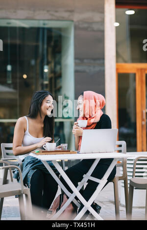 Due happy amici seduti insieme a un cafe' sul marciapiede con il computer portatile Foto Stock