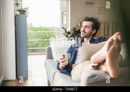 Un giovane seduto sul lettino, utilizzando laptop, bere caffè Foto Stock