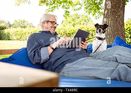 Uomo Senior relax su un letto di oscillazione nel suo giardino, con tavoletta digitale Foto Stock