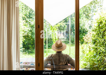 Vista posteriore del senior uomo che indossa cappello di paglia sulla terrazza godendo il suo giardino Foto Stock