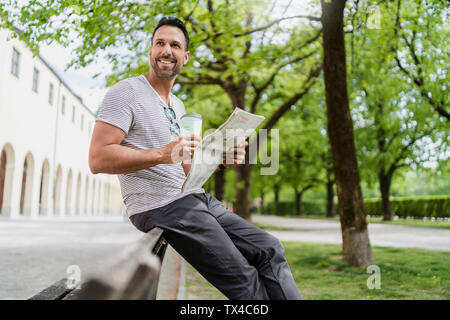 Uomo sorridente con il caffè da asporto e giornale su una panchina nel parco Foto Stock