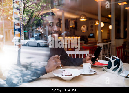 Ritratto di giovane sorridente uomo con gli auricolari e lo smartphone dietro il vetro di finestra in un ristorante Foto Stock