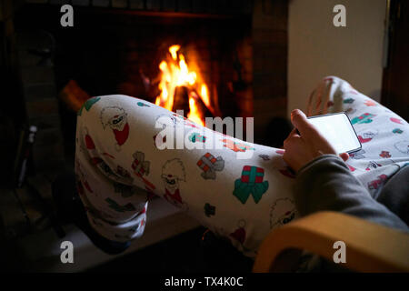 Uomo seduto davanti al camino indossando Babbo Natale Natale pantaloni utilizzando il telefono cellulare Foto Stock