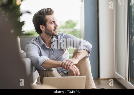Giovane uomo seduto a casa sul pavimento, utilizzando laptop, guardando fuori della finestra Foto Stock