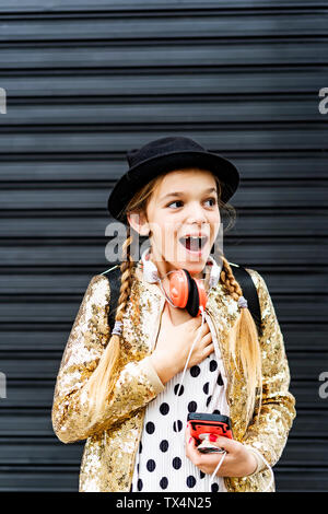 Ritratto di stupito la ragazza con gli auricolari e uno smartphone che indossa un cappello e paillettes dorate camicia Foto Stock