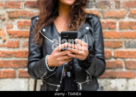 Giovane donna vestita di nero giacca di pelle, utilizza lo smartphone, muro di mattoni in background Foto Stock