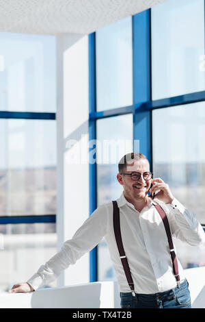 Happy businessman parlando al cellulare nella finestra in ufficio moderno Foto Stock