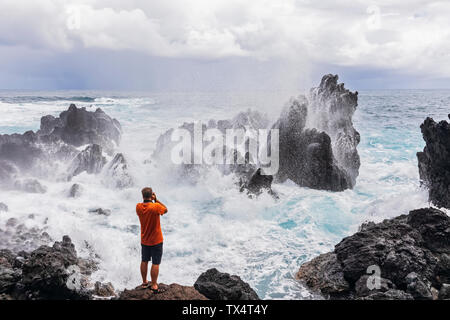 Stati Uniti d'America, Hawaii, Big Island, Laupahoehoe Beach Park,l'uomo a scattare foto di surf di rottura in corrispondenza della costa rocciosa Foto Stock