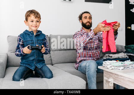 Ritratto di sorridente ragazzino giocare computer gioco mentre suo padre la piegatura servizio lavanderia Foto Stock