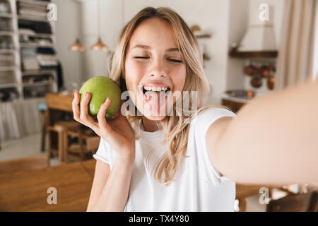 Primo piano immagine di incantevole donna bionda 20s bianco che indossa la t-shirt ridendo e tenendo premuto mela verde tenendo selfie foto in salotto Foto Stock