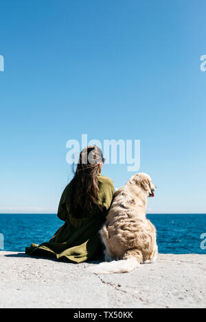 Vista posteriore della donna seduta accanto alla sua Labrador Retriever al quay guardando al mare Foto Stock