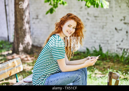 Ritratto di redheaded sorridente giovane donna con lo smartphone seduta sul banco in giardino Foto Stock