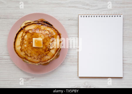 Frittelle fatte in casa con burro e sciroppo d'acero su una piastra di rosa, vuoto notepad bianco su una superficie di legno, vista dall'alto. Il sovraccarico di lavoro piana, laici dal di sopra. Foto Stock