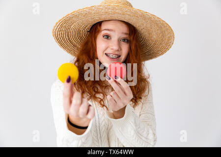 Close up ritratto di un allegro giovane ragazza adolescente che indossa estate hat in piedi isolato su sfondo bianco, tenendo amaretti Foto Stock