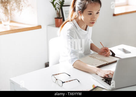 Concentrato asian business donna a scrivere qualcosa e utilizzando il computer portatile mentre seduti a tavola in office Foto Stock