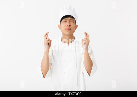 Emozionato chef asiatico che indossano uniformi isolato permanente su sfondo bianco, tenendo le dita incrociate per buona fortuna Foto Stock