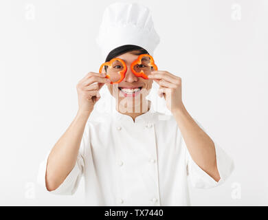 Emozionato chef asiatico che indossano uniformi isolato permanente su sfondo bianco, divertendosi, azienda di capsicum a fette Foto Stock